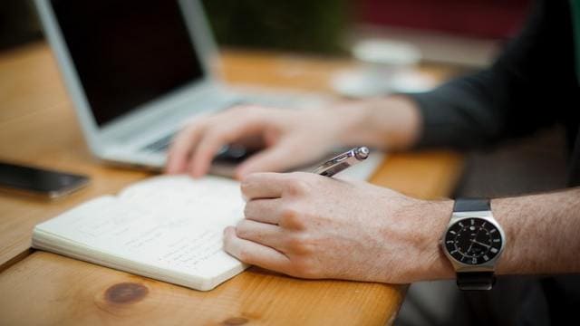 يد رجل تكتب في دفتر بجوار جهاز لابتوب على طاولة خشبية